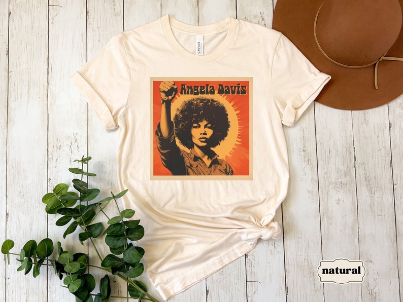 Pu&ograve; includere: Una maglietta beige con una grafica in stile vintage di Angela Davis, una nota attivista, con il testo "Angela Davis" stampato in nero.
