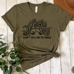 May include: A green t-shirt featuring a black graphic of Medusa's face with snakes for hair. The text "Don't tell me to smile" is printed below the graphic.