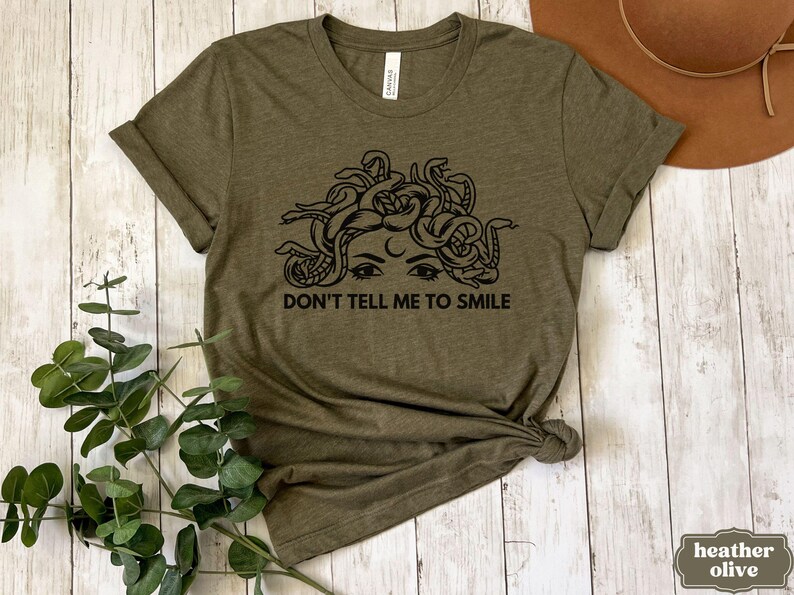 May include: A green t-shirt featuring a black graphic of Medusa's face with snakes for hair. The text "Don't tell me to smile" is printed below the graphic.