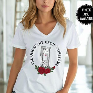 May include: White V-neck t-shirt featuring a graphic of a guillotine surrounded by the text "THE OLIGARCHY GROWS TIRESOME" and red roses. A black and white badge in the upper right corner says "V-NECK ALSO AVAILABLE".