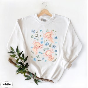 May include: White sweatshirt featuring a colourful print of axolotls and flowers. The axolotls are pink and have blue eyes. The flowers are blue and white.