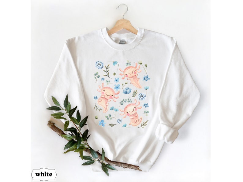 May include: White sweatshirt featuring a colourful print of axolotls and flowers. The axolotls are pink and have blue eyes. The flowers are blue and white.
