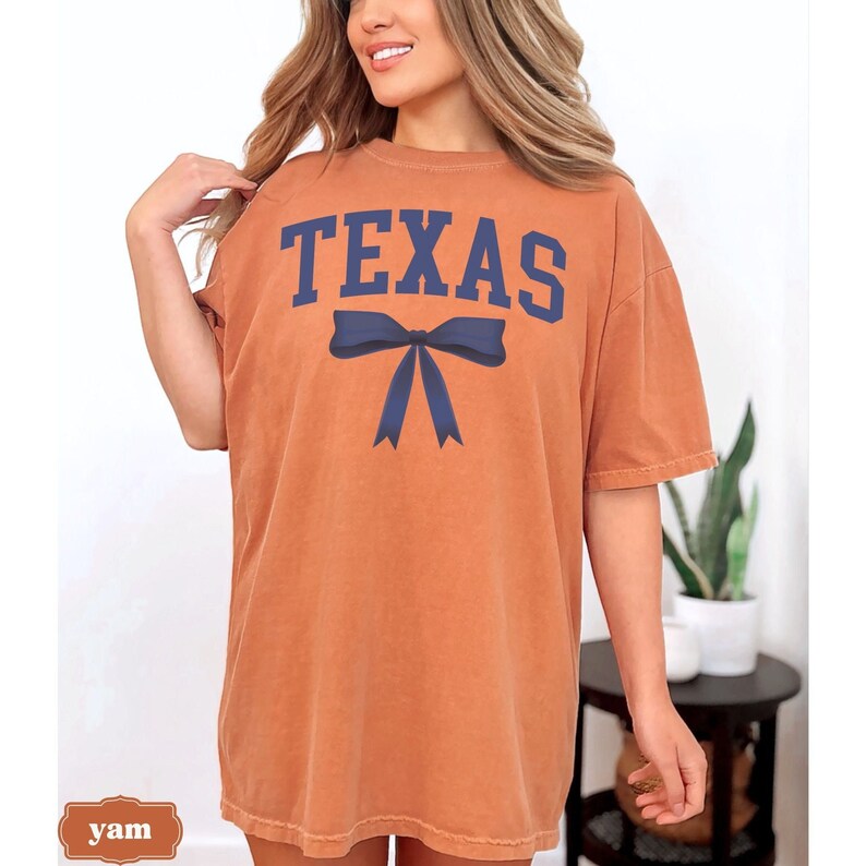 May include: A burnt orange oversized t-shirt with the word "TEXAS" printed in navy blue letters with a navy blue bow underneath.