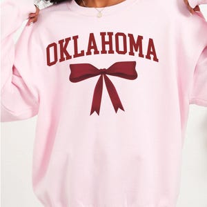 May include: Pink sweatshirt with the word "OKLAHOMA" in maroon letters and a maroon bow.