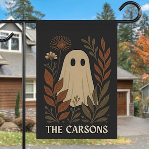 May include: Black garden flag featuring a beige ghost illustration surrounded by autumn-colored leaves and flowers. The text "THE CARSONS" is displayed at the bottom in white. A decorative outdoor accent for Halloween.