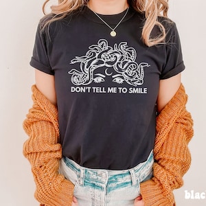 K&ouml;nnte beinhalten: Schwarzes T-Shirt mit einem wei&szlig;en Grafikdruck eines Frauenkopfes mit Schlangen als Haaren. Unter dem Grafikdruck steht der Text "Don't tell me to smile."