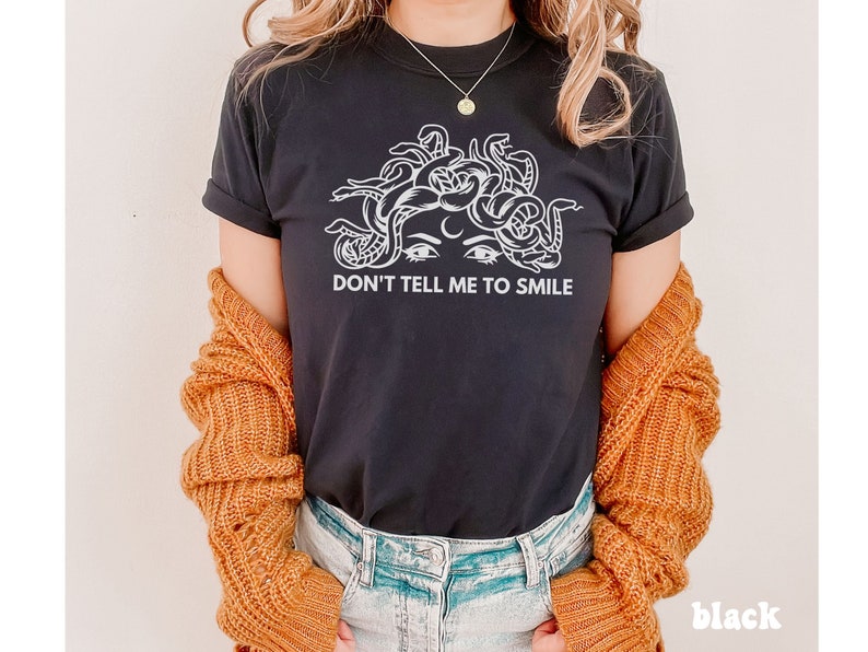 K&ouml;nnte beinhalten: Schwarzes T-Shirt mit einem wei&szlig;en Grafikdruck eines Frauenkopfes mit Schlangen als Haaren. Unter dem Grafikdruck steht der Text "Don't tell me to smile."