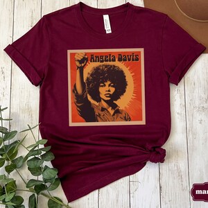 Pu&ograve; includere: Maglietta color marsala con una grafica in stile vintage di Angela Davis con il testo "Angela Davis" in uno stendardo giallo.