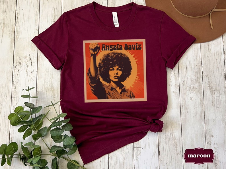 Pu&ograve; includere: Maglietta color marsala con una grafica in stile vintage di Angela Davis con il testo "Angela Davis" in uno stendardo giallo.