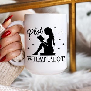 Puede incluir: Taza de cerámica blanca con el texto "Plot WHAT PLOT" y una silueta negra de una persona leyendo. El diseño incluye estrellas. La taza la sostiene una persona que lleva un jersey color crema.