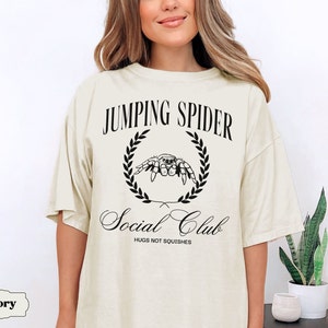 May include: A light ivory t-shirt with a black graphic of a jumping spider in a laurel wreath. The text "Jumping Spider Social Club" is printed below the spider, with the words "Hugs not squishes" printed below the text.