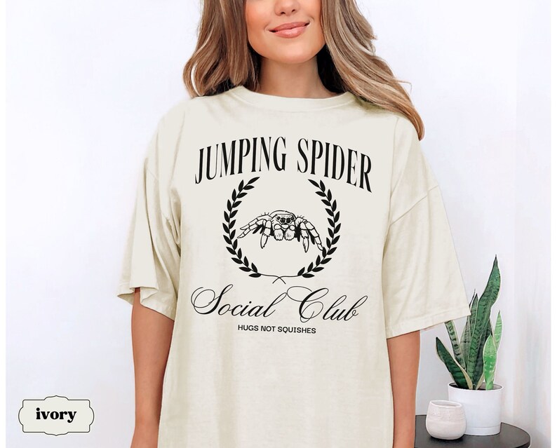 May include: A light ivory t-shirt with a black graphic of a jumping spider in a laurel wreath. The text "Jumping Spider Social Club" is printed below the spider, with the words "Hugs not squishes" printed below the text.