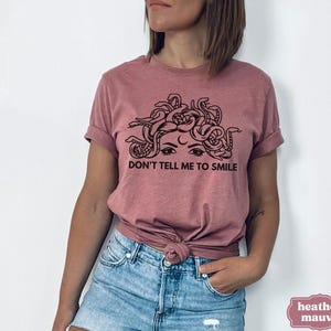 May include: A heather mauve t-shirt featuring a black and white graphic of a woman's face with snakes for hair. The text "Don't tell me to smile" is printed below the graphic.