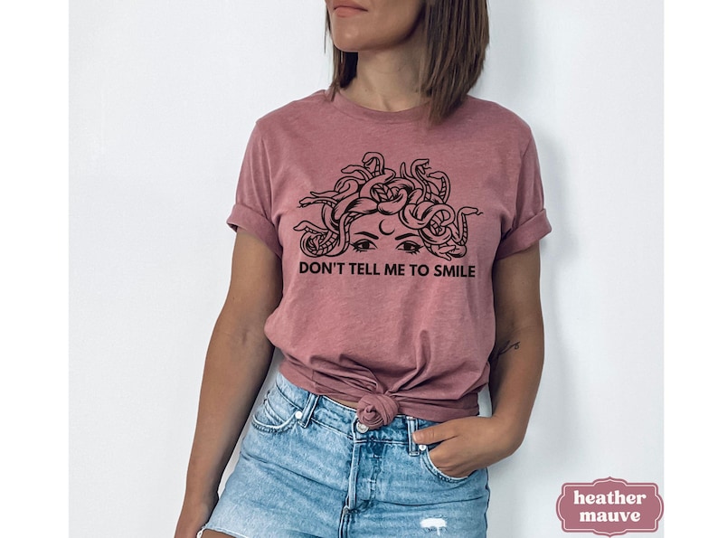 May include: A heather mauve t-shirt featuring a black and white graphic of a woman's face with snakes for hair. The text "Don't tell me to smile" is printed below the graphic.