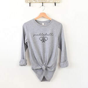 May include: A heather gray long-sleeve shirt with the word "pickleball" in a script font above a graphic of two crossed pickleball paddles and a ball. The shirt is knotted at the waist.