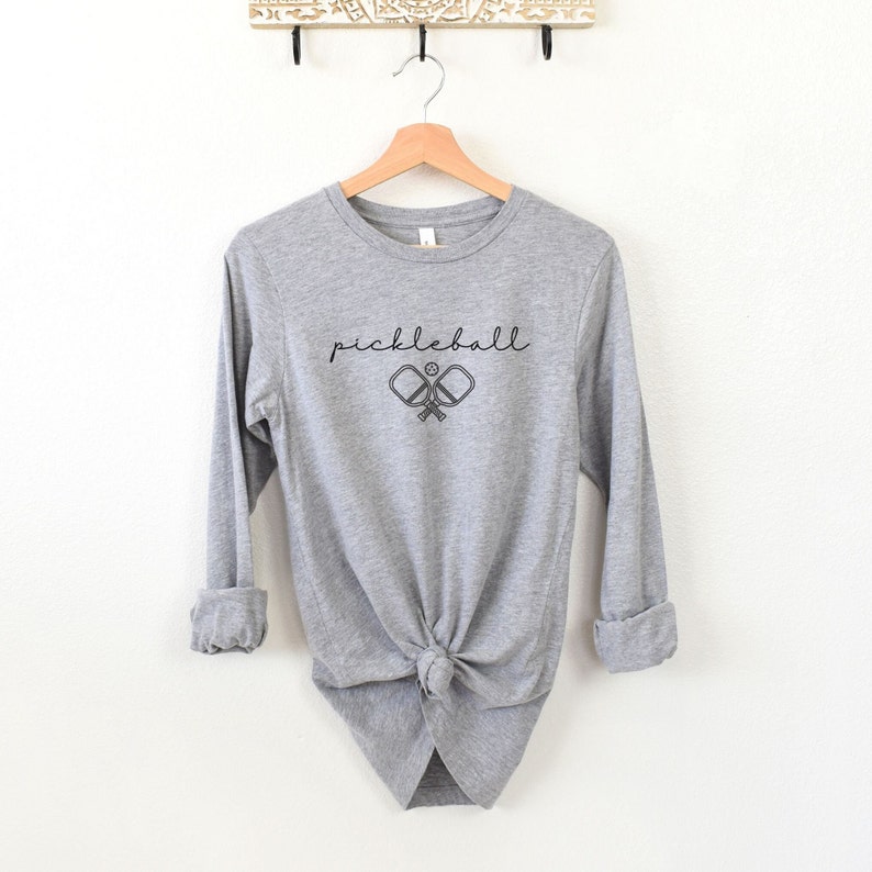 May include: A heather gray long-sleeve shirt with the word "pickleball" in a script font above a graphic of two crossed pickleball paddles and a ball. The shirt is knotted at the waist.