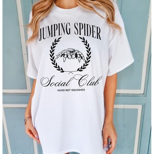 May include: White oversized t-shirt with black text that reads "Jumping Spider Social Club" and a black illustration of a spider in a wreath. The text below the spider reads "Hugs not squishes."