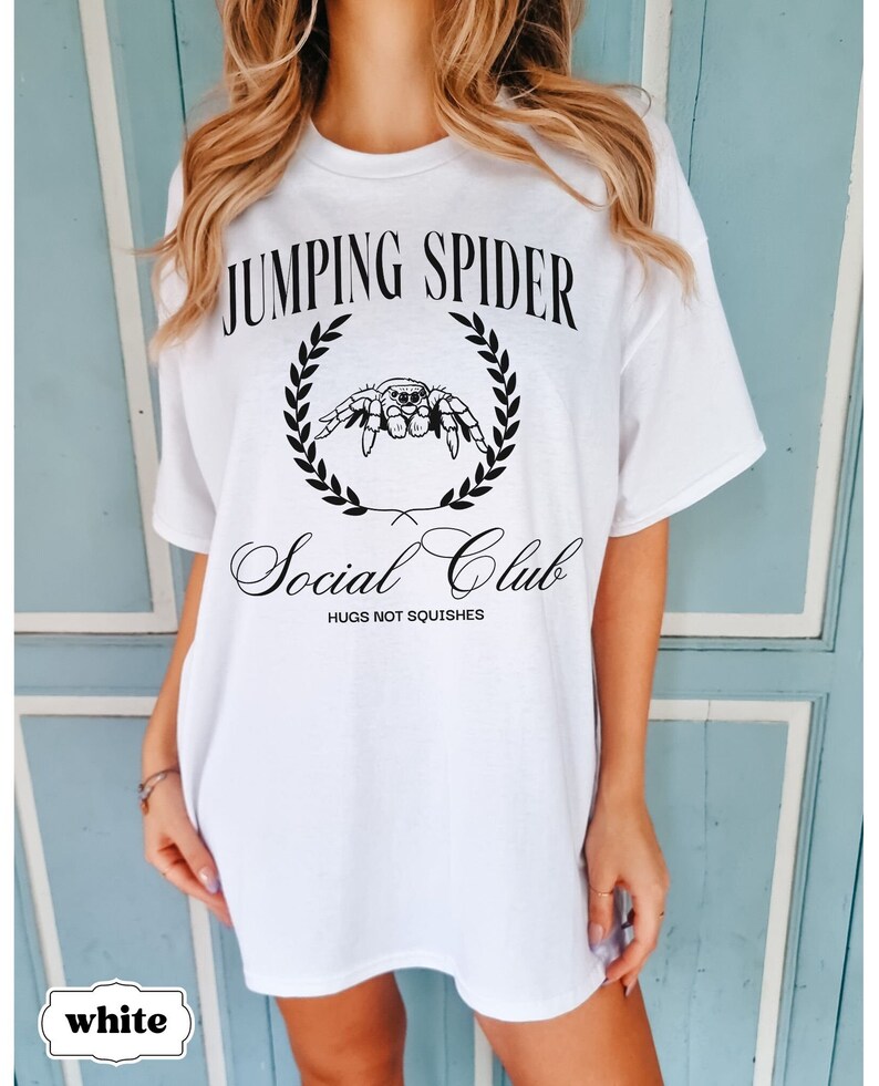 May include: White oversized t-shirt with black text that reads "Jumping Spider Social Club" and a black illustration of a spider in a wreath. The text below the spider reads "Hugs not squishes."