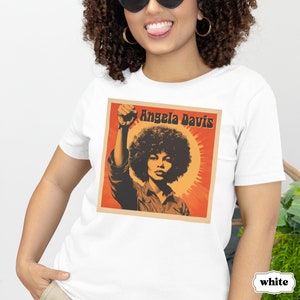 Pu&ograve; includere: Maglietta bianca con una grafica di Angela Davis con il pugno alzato. La grafica &egrave; in stile retr&ograve; con colori arancio, rosso e giallo. Il testo "Angela Davis" &egrave; stampato in nero sopra la grafica.