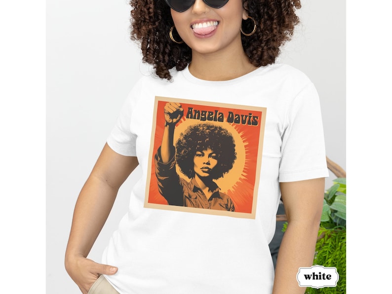 Pu&ograve; includere: Maglietta bianca con una grafica di Angela Davis con il pugno alzato. La grafica &egrave; in stile retr&ograve; con colori arancio, rosso e giallo. Il testo "Angela Davis" &egrave; stampato in nero sopra la grafica.