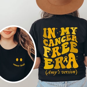 Puede incluir: Camiseta negra con el texto "IN MY CANCER FREE ERA (Amy's version)" en letras amarillas de estilo retro. La parte posterior de la camiseta presenta una pequeña cinta amarilla.