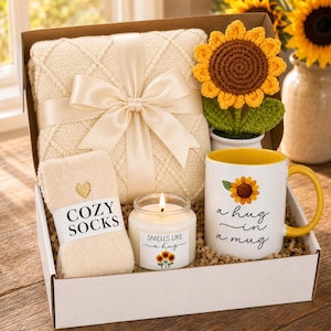May include: A gift box containing a cream-colored textured blanket tied with a satin ribbon, a crocheted sunflower in a white pot, a pair of cream-colored socks labeled COZY SOCKS, and a white mug with a yellow interior and the text A hug in a mug.