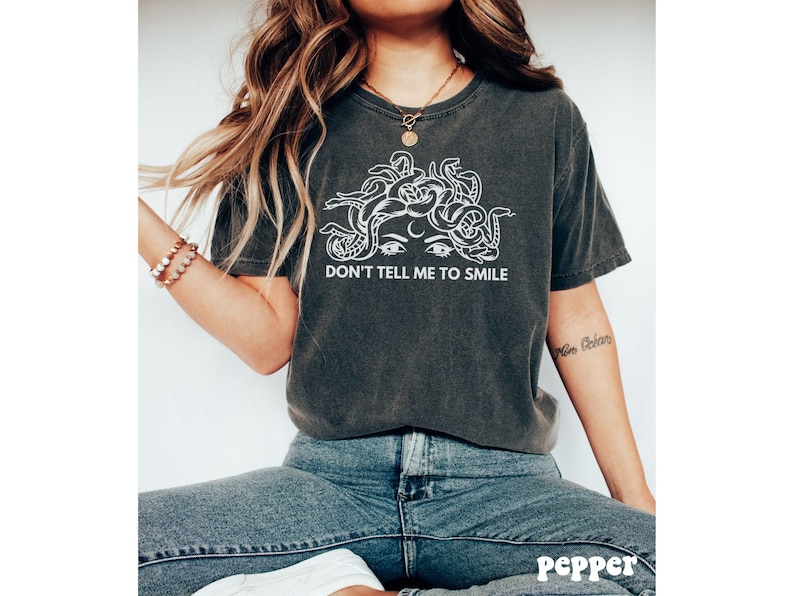 K&ouml;nnte beinhalten: Ein dunkelgraues T-Shirt mit einem wei&szlig;en Motiv von Medusas Gesicht mit Schlangen als Haaren. Der Text "Don't Tell Me To Smile" ist unter dem Motiv gedruckt.