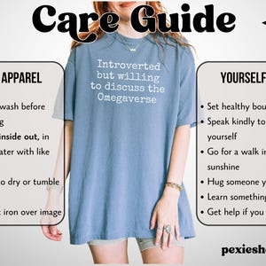May include: A pale blue t-shirt with the white text "Introverted but willing to discuss the Omegaverse". The image also includes a care guide with washing instructions and self-care tips.