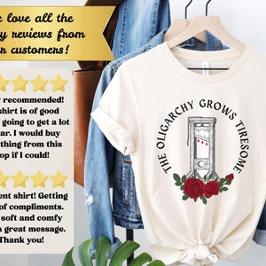 May include: Cream-colored t-shirt featuring a guillotine graphic with red roses and the text "THE OLIGARCHY GROWS TIRESOME." The shirt is hanging on a wooden hanger.