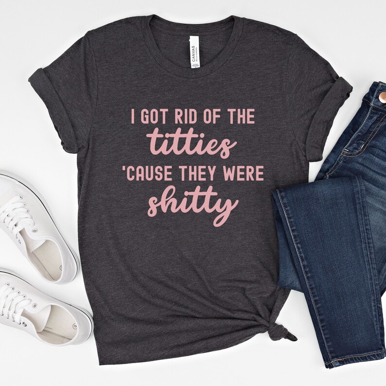 Op de afbeelding: Donkergrijs t-shirt met de roze tekst "I GOT RID OF THE titties 'CAUSE THEY WERE shitty". Het shirt is gestyled met een blauwe jeans en witte sneakers.