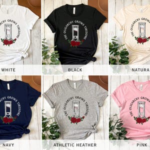 May include: A collection of t-shirts in various colors, including white, black, navy, and pink. Each shirt features a guillotine graphic with the text "THE OLIGARCHY GROWS TIRESOME" in a circular design, surrounded by red roses.