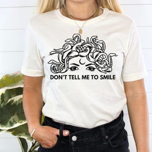 May include: White t-shirt featuring a black and white illustration of a woman with snakes for hair. The text "Don't tell me to smile" is printed below the illustration.