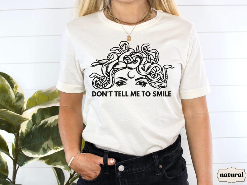 May include: White t-shirt featuring a black and white illustration of a woman with snakes for hair. The text "Don't tell me to smile" is printed below the illustration.