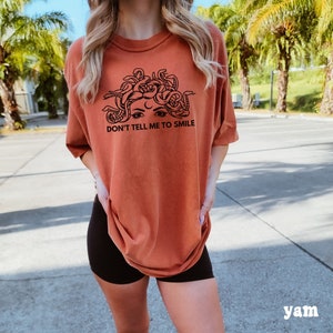 K&ouml;nnte beinhalten: Ein rostfarbenes T-Shirt mit einem schwarzen Motiv von Medusas Gesicht mit Schlangen als Haaren. Der Text "Don't Tell Me To Smile" ist unter dem Motiv gedruckt.