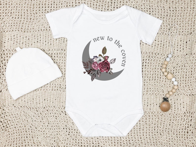 K&ouml;nnte beinhalten: Wei&szlig;er Baby-Strampler mit einem grauen Halbmond und einem Blumendesign. Der Text "new to the coven" ist auf dem Strampler aufgedruckt.