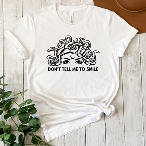 May include: White t-shirt featuring a black and white illustration of a woman with snakes for hair. The text "Don't tell me to smile" is printed below the illustration.
