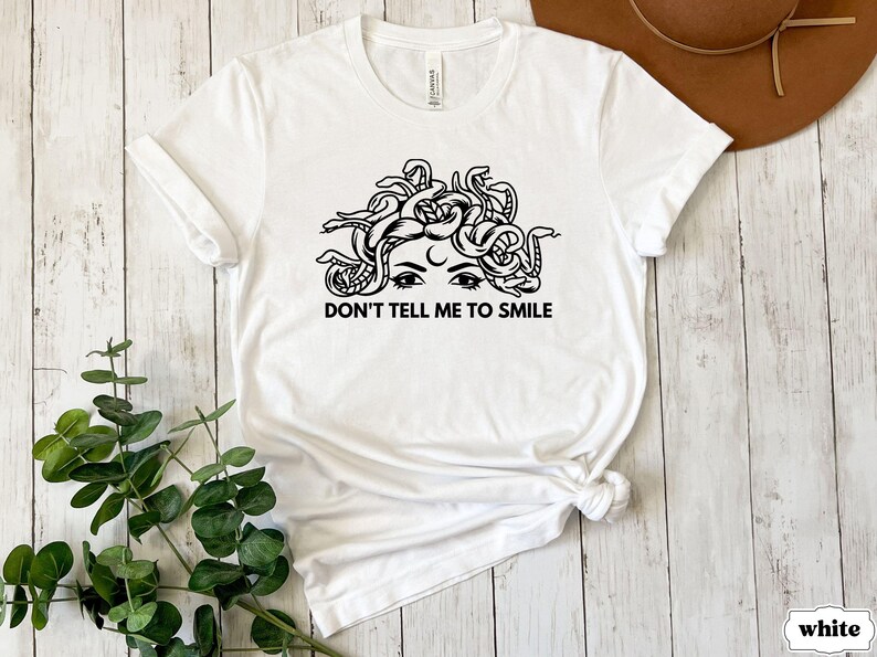 May include: White t-shirt featuring a black and white illustration of a woman with snakes for hair. The text "Don't tell me to smile" is printed below the illustration.