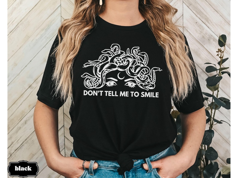 May include: Black t-shirt featuring a white graphic of Medusa's head with snakes and the text "Don't tell me to smile".
