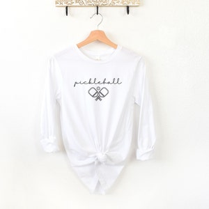 May include: White long-sleeve shirt with the word "pickleball" in a script font above a graphic of two crossed pickleball paddles and a ball. The shirt is hanging on a wooden hanger.