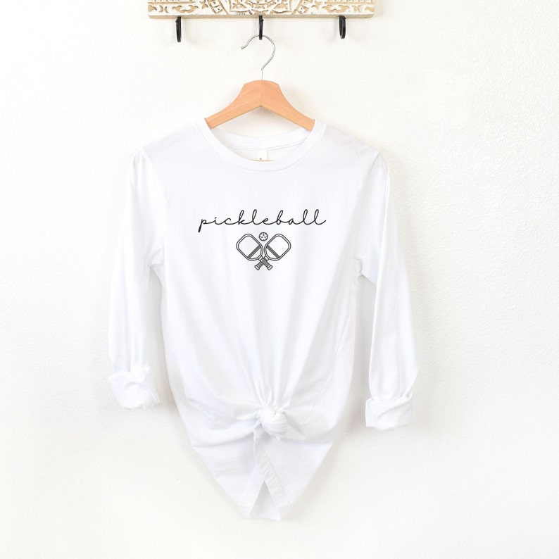 May include: White long-sleeve shirt with the word "pickleball" in a script font above a graphic of two crossed pickleball paddles and a ball. The shirt is hanging on a wooden hanger.