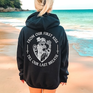 May include: Black hooded sweatshirt with a white graphic print of two skeletons holding a rose in a heart shape. The text reads "From our first kiss till our last breath".