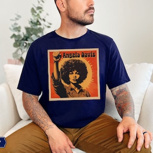 Pu&ograve; includere: Una t-shirt blu navy con una grafica in stile vintage di Angela Davis con il testo "Angela Davis" in bianco.