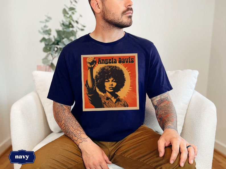 Pu&ograve; includere: Una t-shirt blu navy con una grafica in stile vintage di Angela Davis con il testo "Angela Davis" in bianco.