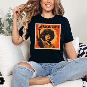 Pu&ograve; includere: Una maglietta nera con una grafica in stile vintage di Angela Davis con il testo "Angela Davis" stampato sul davanti.