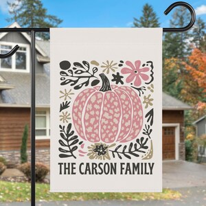 Puede incluir: Una bandera de jardín crema con un diseño de calabaza rosa rodeada de ilustraciones florales y de hojas en negro, dorado y rosa. El texto "THE CARSON FAMILY" está impreso en negro en la parte inferior.