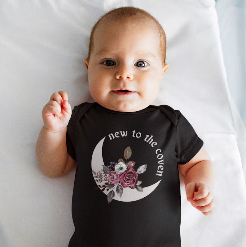 K&ouml;nnte beinhalten: Ein schwarzer Baby-Strampler mit einem wei&szlig;en Halbmond und einem Blumendesign. Der Text "new to the coven" ist um den Mond geschrieben.