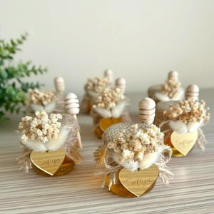 May include: Small glass jars filled with honey, adorned with dried flowers, burlap, and wooden honey dippers. Each jar has a heart-shaped tag with the name "Safiya" engraved on it. A charming gift idea.