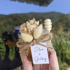 May include: A small glass jar filled with honey, adorned with dried flowers, fluffy accents, and a wooden honey dipper. A tag tied with twine reads "Zilan & Gulbey". The jar is held in front of a blurred outdoor background.