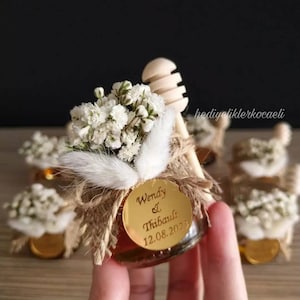 May include: Small glass jar filled with honey, decorated with burlap, white flowers, and a wooden honey dipper. A gold round label with the text "Wendy & Thibault 12.08.2023" is attached to the jar.