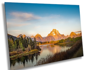 Puede incluir: Una vista panorámica de una cordillera con un lago en primer plano. Las montañas son doradas con picos cubiertos de nieve. El lago está tranquilo y refleja las montañas y el cielo. El cielo es azul claro con algunas nubes blancas.